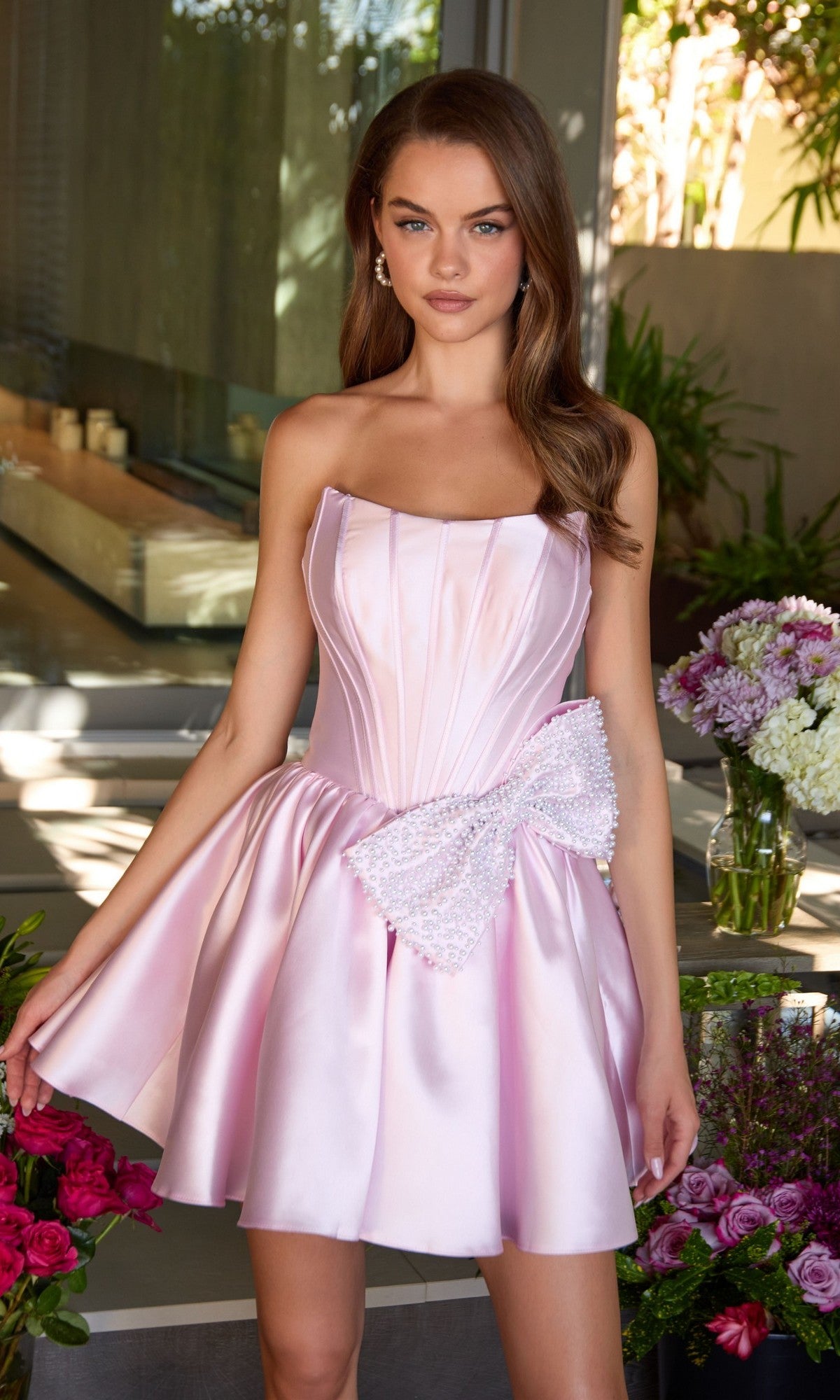 A woman in the Short Homecoming Dress: Ava Presley 47298, featuring a strapless satin design with a boned corset bodice and beaded bow, stands indoors by floral arrangements.
